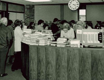 group of people lined up around a library circulation desk with several large stacks of books on the top