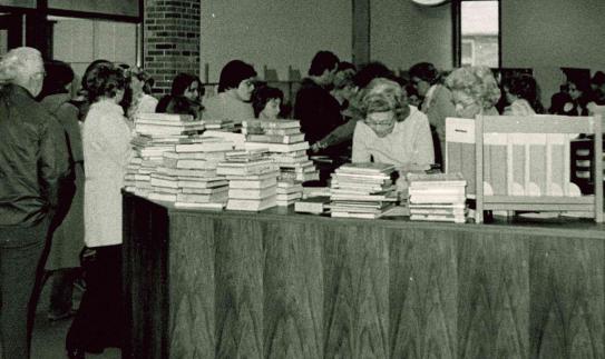 group of people lined up around a library circulation desk with several large stacks of books on the top